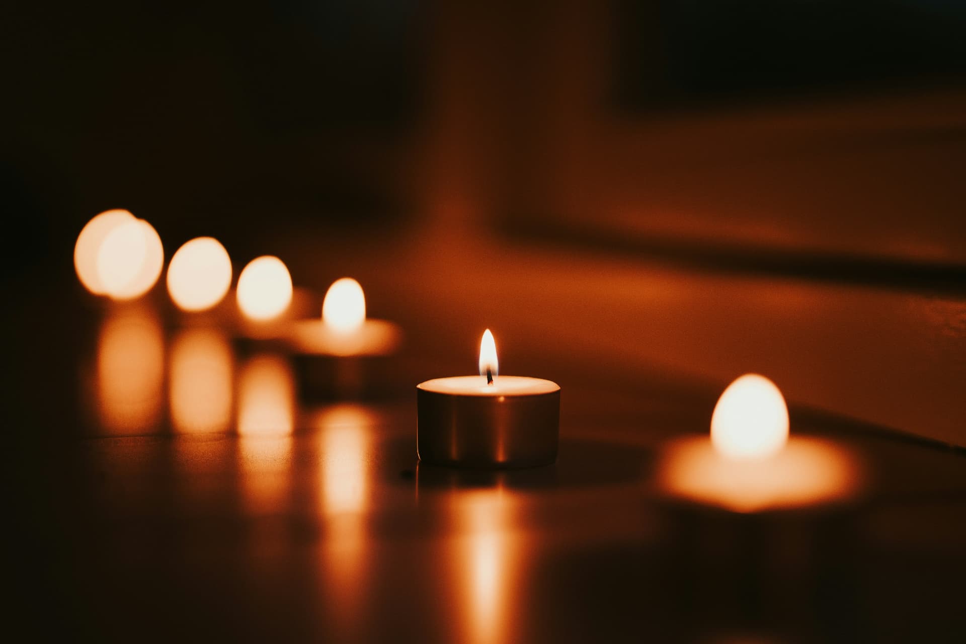 A line of small lit tealights glowing in the dark, their flames mirrored in a polished surface